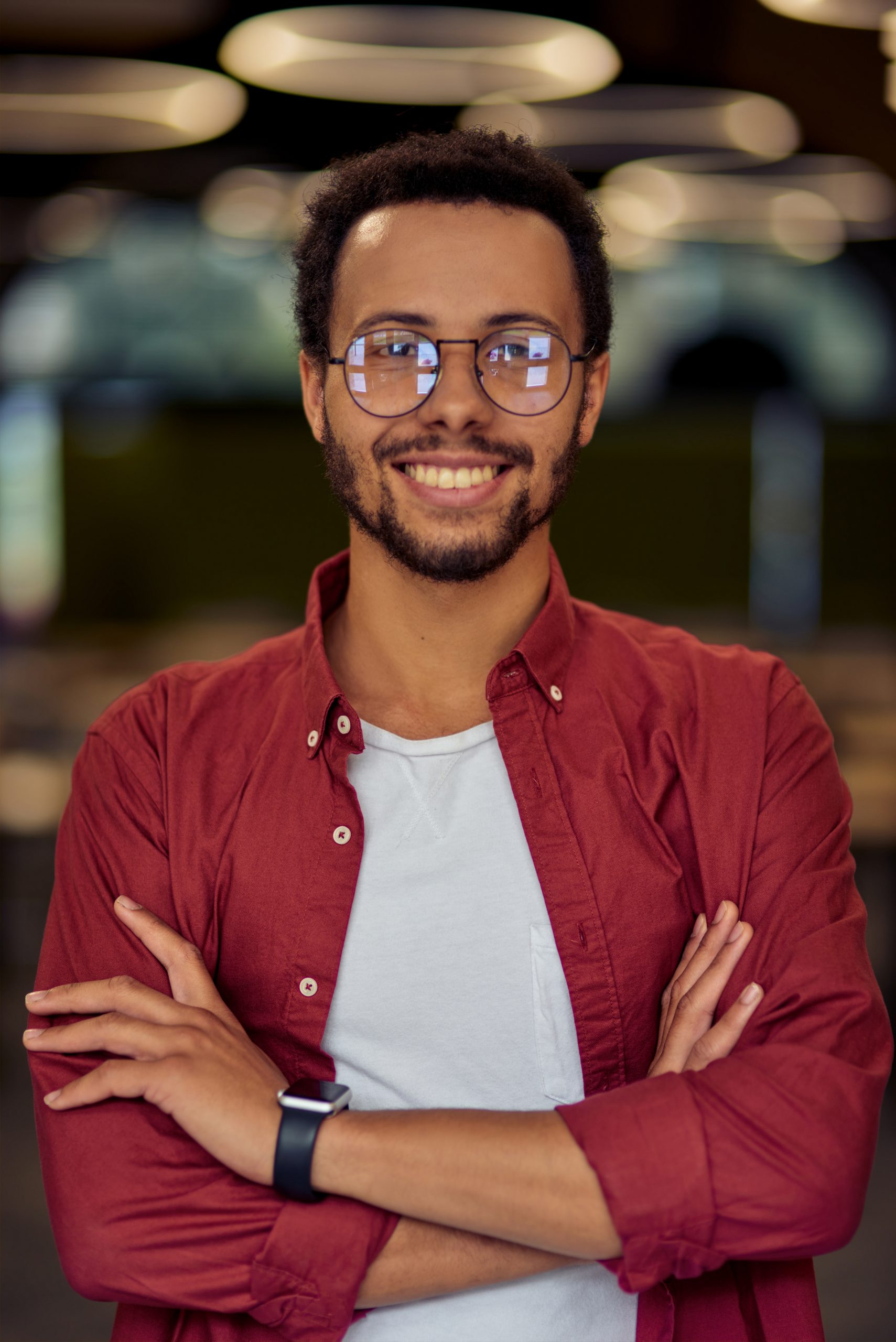 Vertical shot of a young happy mixed race businessman or office worker in casual wear keeping arms crossed, looking at camera and smiling while posing in the office