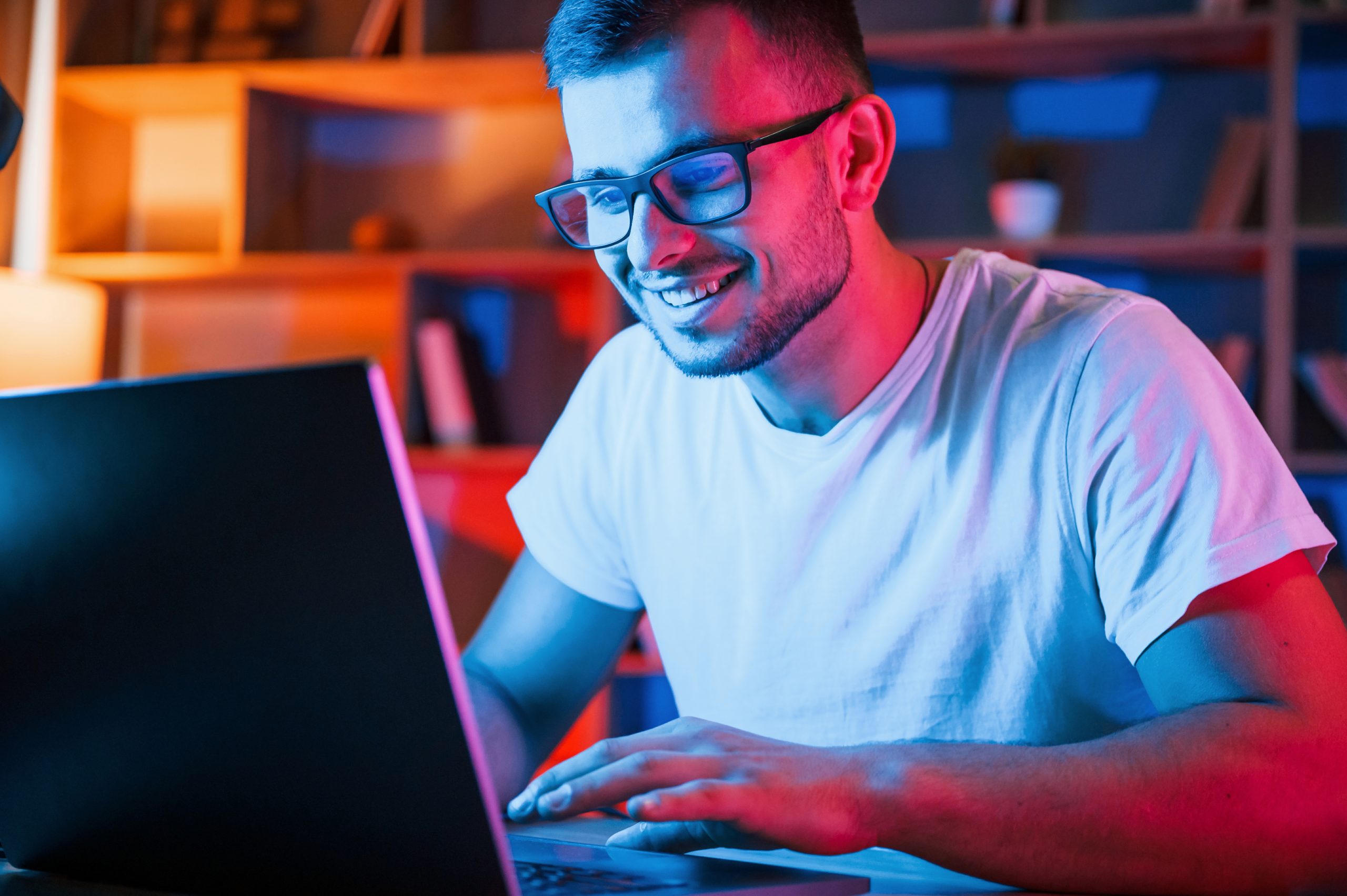 Positive emotions. Man in glasses and white shirt is sitting by the laptop in dark room with neon lighting